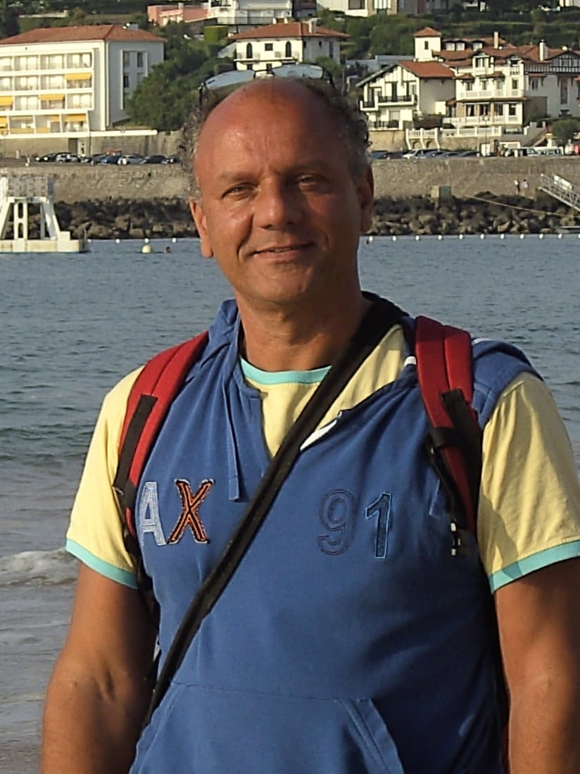 Esam Abou-El Magd steht lächelnd am Strand vor dem Meer, die Trägergurte eines Rucksacks über den Schultern; im Hintergrund die Bucht und Häuser am Hang.